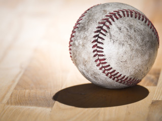 Close up sports background image of an old used weathered leather baseball showing intricate detailing and red laces sitting on a wood butcher block counter with lighting from behind and dark shadows.