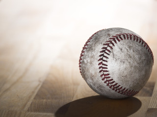 Close up sports background image of an old used weathered leather baseball showing intricate detailing and red laces sitting on a wood butcher block counter with lighting from behind and dark shadows.