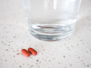Close up photograph of two red or brown oblong shaped pills or medicine laying next to a glass of water on a white quartz countertop making a great medical or health care related background.