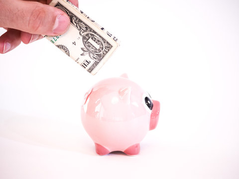 Photograph of a Caucasian male hand holding a United States dollar bill folded up and inserting it in a pink ceramic piggy bank sitting on a table isolated on a white background.