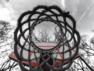 Close up unique photograph view looking up at an urban or city playground basketball net and hoop with the sky, clouds and tree branches above and orange and white backboard.
