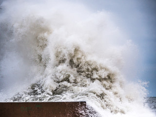 Photograph of a large wave from Lake Michigan crashing into concrete wall barrier and splashing and spilling over the edge with blue sky beyond making a dramatic background photograph. - Powered by Adobe