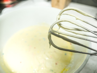 Close up of mixed yellow colored cake batter with sprinkles and metal beaters with mixture dripping from the tips on top of a white kitchen countertop.