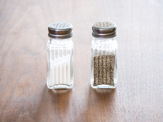 Close up view of a glass and metal salt and a pepper shaker about half full of seasoning sitting on a dark wood grain table top with fluted square shaped container and rounded silver colored tops.