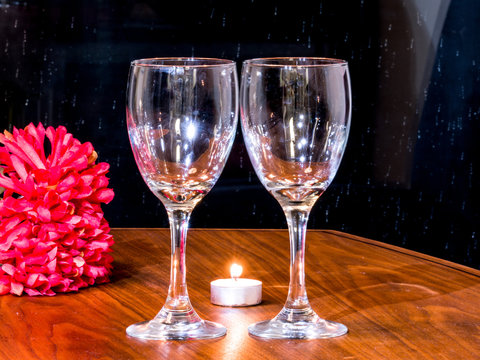 Romantic background photograph of two empty wine glasses displayed on a dark wood grain table with tea candles and red flowers in the background.