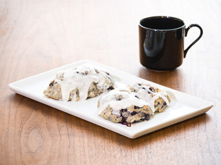 Two blueberry scones sitting on a white serving plate next to a black porcelain coffee or tea mug on a dark grained dining table making for a beautiful breakfast or tea time background.
