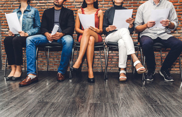 Group of business people holding paper while sitting on chair waiting for job interview against brick background - obrazy, fototapety, plakaty