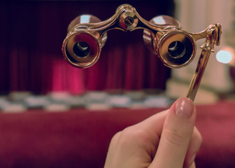 Woman hand holds opera glasses in theater against stage and drop-curtain - obrazy, fototapety, plakaty