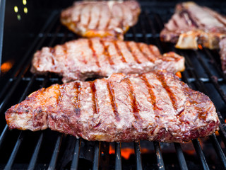 Close-up photograph of various cuts of beef including filet mignon and t-bone steak on a gas fire grill for a backyard barbeque.