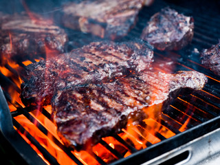 Close-up photograph of various cuts of beef including filet mignon and t-bone steak on a gas fire grill for a backyard barbeque.