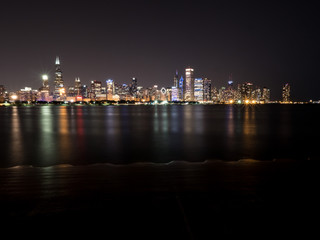 Fototapeta premium Beautiful long exposure Chicago night skyline photo with colorful red, green, purple, blue, pink, orange, and yellow building lights and water reflections on Lake Michigan and shoreline in foreground