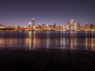 Fototapeta premium Beautiful long exposure Chicago night skyline photo with colorful red, green, purple, blue, pink, orange, and yellow building lights and water reflections on Lake Michigan and shoreline in foreground
