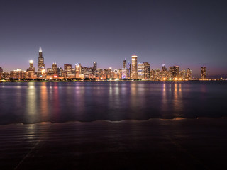 Fototapeta na wymiar Beautiful long exposure Chicago night skyline photo with colorful red, green, purple, blue, pink, orange, and yellow building lights and water reflections on Lake Michigan and shoreline in foreground