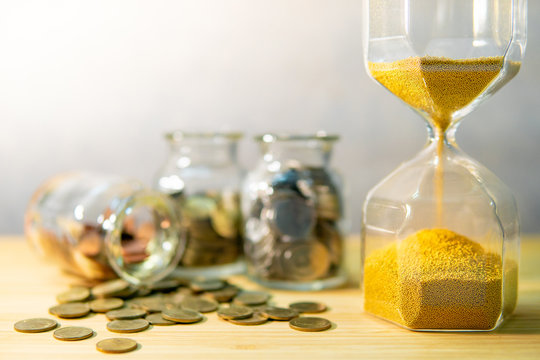 Sand running through the shape of hourglass with coins in currency glass jars on wooden table. Saving money for future retirement. Time investment concept