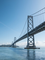 Gorgeous photograph of the famous and expensive San Francisco - Oakland Bay Bridge from the edge of the water looking across the bay with blue sky above.