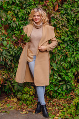 Young beautiful woman in sweater and coat stands next to the background of wild grapes holding bouquet of autumn leaves