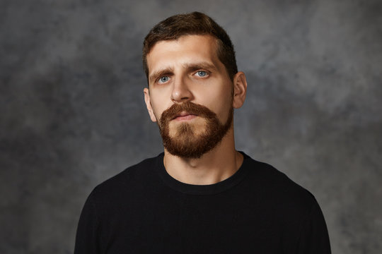Isolated shot of serious handsome thirty year old Caucasian man with blue eyes, mustache and thick beard posing against blank studio wall background, looking at camera, dressed in black sweater