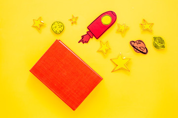 Literature for children. Fantactic, fiction story. Book with blank cover near cutout of rocket, stars, moon on yellow background top view mockup copy space