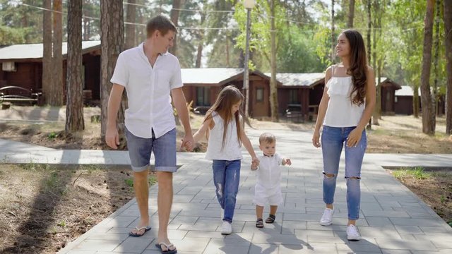 happy family is walking on resort area in forest in summer day, holding hands of their little children