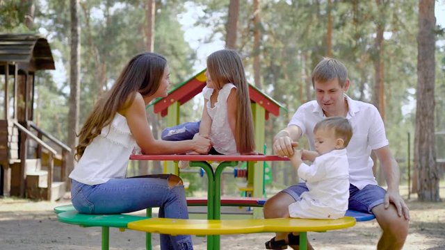 happy parents with their little children are resting on playground in park with high spruce in summer day