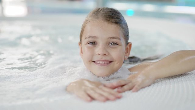 pretty little girl with wet hair is sitting alone in hydromassage bath in a water park, looking to camera