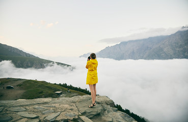 Obraz premium A young Girl in a yellow raincoat photographs the mountains. Georgia. Summer. August. Girl making a photo shoot of mountain. 