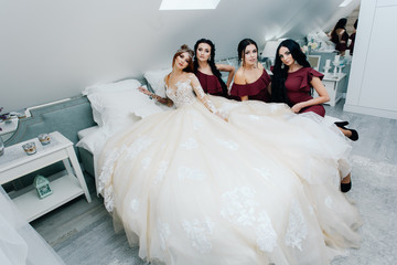 the red-haired bride sits on the bed in the morning with the bridesmaids in marsala dresses