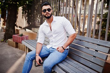 Stylish tall arabian man model in white shirt, jeans and sunglasses posed at street of city. Beard attractive arab guy sitting on bench.