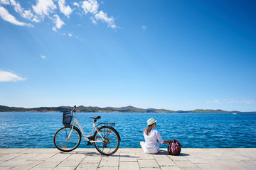 Back view of young woman in white closing, sunhat and sunglasses sitting at bicycle on stony sidewalk under clear blue sky on sparkling clear water background. Tourism and vacations concept. - Powered by Adobe