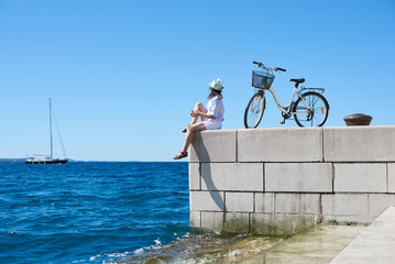 Back view young tourist woman in white clothing and sunglasses sitting at bicycle on high paved...