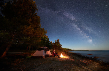 Night camping at sea coast. Female hiker sitting relaxed in front of tent at campfire under bright starry sky and Milky way, enjoying beautiful view of blue water. Tourism and active lifestyle concept