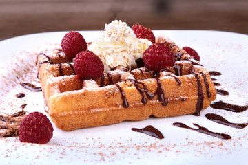 Closeup of homemade Belgian waffles with whipped cream, chocolate sauce and raspberries on white plate.