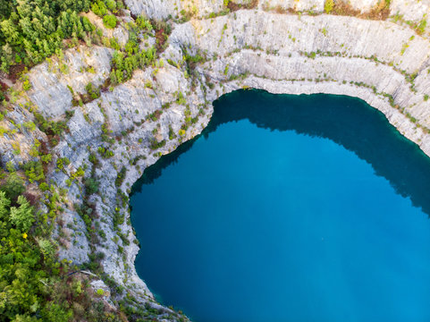 (Laufaufnahme, Drohne) Felswand eines Steinbruchs mit blauem See im Vordergrund
