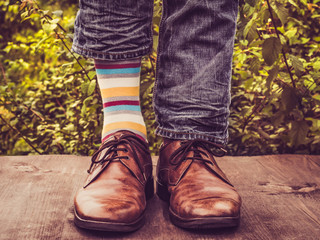 Men's legs in stylish shoes, bright, multi colored, variegated socks with Christmas and New Year's patterns on the wooden terrace on the background of green trees. Beauty, fashion, elegance