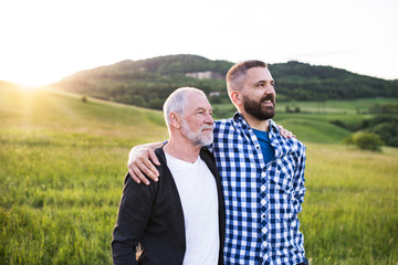 A portrait of an adult hipster son with senior father in nature at sunset, arms around each other.