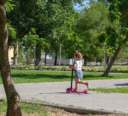 Fototapeta premium Cute child skilfully skates on a pink two-wheeled scooter on a summer sunny day