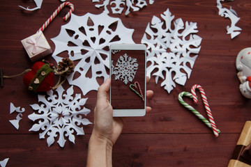 New Year's Christmas concept. Women's hands hold a smartphone, photograph snowflakes cut from paper, gifts, scissors on a wooden table