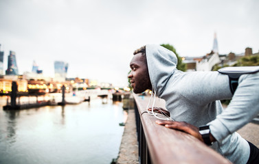 Black man runner with smartphone in an armband on the bridge in a city, resting.