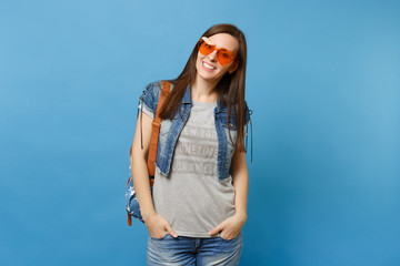 Young cheerful woman student in denim clothes with backpack wearing orange heart glasses keeping hands in pockets isolated on blue background. Education in university. Copy space for advertisement.
