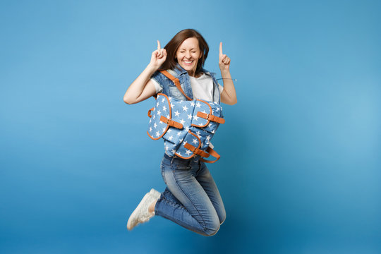 Full length portrait young overjoyed woman student with closed eyes with backpack jumping pointing index figers up isolated on blue background. Education in high school. Copy space for advertisement.