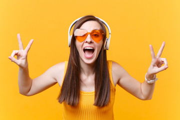 Portrait of excited fun girl in orange glasses listening music in headphones showing victory...