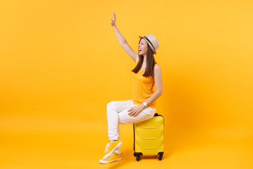 Fototapeta na wymiar Tourist woman in summer hat sit on suitcase waving and greeting with hand as notices someone isolated on yellow background. Passenger traveling abroad to travel on weekends getaway. Air flight concept