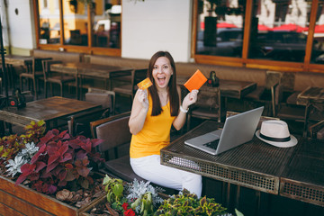 Woman in outdoors street cafe sitting at table with modern laptop pc computer, hold in hand bank...
