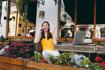 Happy girl in outdoors street cafe sitting at table with laptop pc computer, talking on mobile phone, conducting pleasant conversation, in restaurant during free time. Mobile office freelance concept
