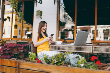 Happy surprised girl in outdoors street coffee shop cafe sitting at table with laptop computer, texting message on mobile phone friend, in restaurant during free time. Mobile office freelance concept