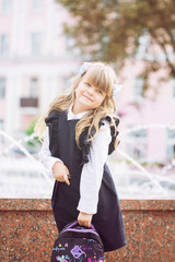 A small cheerful blue-eyed girl, a first-class woman with winded hair with bows and glasses in school uniform with a briefcase and book on September 1, is sitting in the school park near the fountain