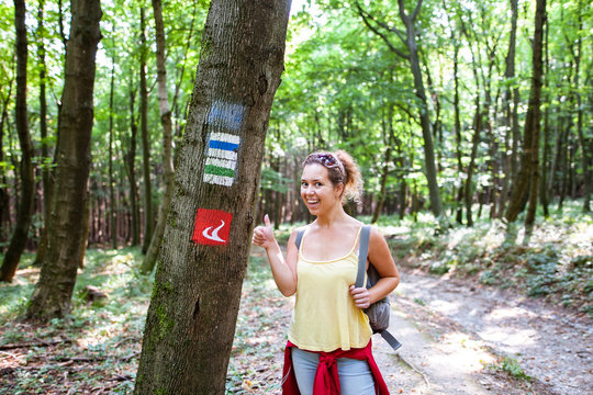 Beautiful woman smiling at path in a forest nex to a tourist sign during hiking on a hill during sunrise on summer