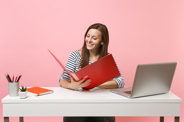 Successful woman looking on red folder with paper documents working on project while sitting at office with laptop isolated on pastel pink background. Achievement business career concept. Copy space.