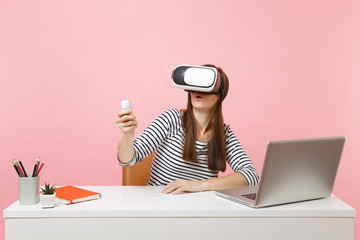 Young amazed woman in headset of virtual reality on head using remote control sit work at white desk with pc laptop isolated on pastel pink background. Achievement business career concept. Copy space.