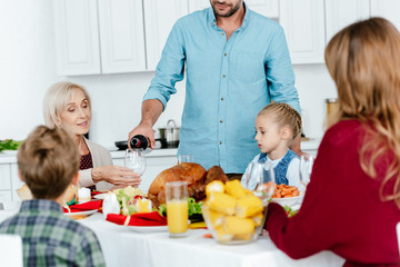 partial view of man pouring wine into glass while family celebrating thanksgiving at served table with baked turkey